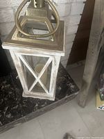 White wooden lantern with glass panels, metal top and handle, and candle inside, displayed on black granite surface near a fireplace and wooden decoration.