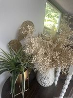 Photo showing large white vase with artificial off-white foliage, two woven chairs, and green leafy plant in gold planter