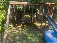 Full view of wooden kids swing set with two yellow belt swings, yellow trapeze bar, and blue slide attached on right.