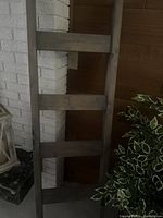 Wooden ladder leaning against a white brick wall beside a plant, showing four wide rungs and rustic wood finish