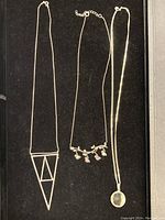 Full view of three silver necklaces laid on black background showing different pendants: geometric triangular shape, bird branch, and round faceted pendant.