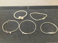 Five silver bracelets laid out on black surface showing varied styles including beaded, chain, bangles, and open cuff designs.