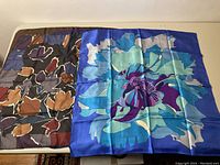 Two folded silk scarves showing different designs and colors, including a floral blue and purple and an abstract brown and black.