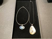 Two necklaces with pendants displayed on black velvet surface. Left: Sterling silver chain with oval iridescent pendant. Right: Black leather rope necklace with teardrop-shaped shell pendant with pearl detail.