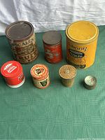 Photo showing seven vintage tins of varying sizes, colors and designs, displayed on a green cloth background.