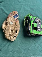 Photo showing two baseball gloves side by side on a green table cover, one tan well-used Spalding 11-inch glove and one black and green Rawlings 9-inch glove.