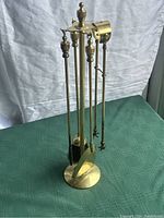 Full view of brass fireplace tools set with brush, poker, shovel, tongs and brass stand on green surface and white draped backdrop.