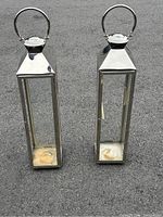 Two tall, narrow stainless steel candle holders, missing glass, showing front and top view with handles and hinged lids.