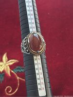 Close-up image of men's 925 sterling silver ring mounted on a ring sizing tool showing size 12 with a prominent oval red stone and detailed silver filigree band.