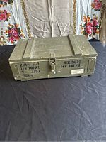 Front view of a rectangular vintage olive green wooden box with metal latch and stenciled markings, placed on black cloth with floral backdrop.