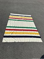 Top view of cream colored wool blanket with green, red, yellow, and navy blue stripes laid flat on blacktop.