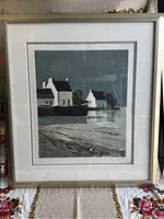 Framed lithograph print showing a scene with houses near water, with visible signature and numbering at bottom, displayed against a floral tablecloth.