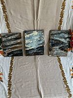 Photo showing three small vintage oil paintings on board, all featuring dark rain-themed abstract scenes with a mix of black, white, gray, and brown colors, laid out on a beige cloth with decorative borders.