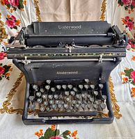 Front and angled top view of vintage Underwood Standart typewriter showing keyboard and paper feed mechanism against floral backdrop.