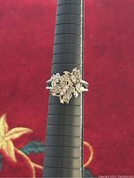 Front view of a silver floral cluster ring placed on a black ring mandrel against a red fabric background. The floral design features multiple flowers with small stones embedded.