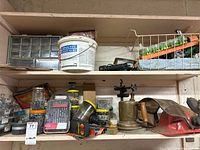 Two wooden shelves with assorted hardware items including a container of joint sealer, hardware organizer, drill bits and nails, blow torch, red dustpan, and hardware boxes.