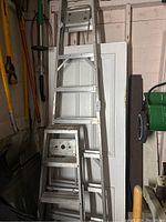 3 aluminum ladders including step and extension types stacked against wall with tools around.