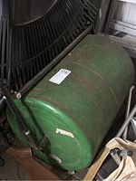 Green lawn roller with metal drum and black handle showing rust and use signs, positioned indoors leaning against a wall and surrounded by other garden equipment.
