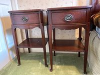 Front view of two matching wooden side tables with drawers and lower shelves placed side by side on green carpet.
