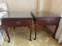 Two matching wooden side tables with glass top protectors and decorative drawer handles, placed side by side against a wall.