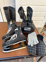 Full view of the lot showing all items - Bauer skates, rubber boots, winter boots, slippers, gloves, and hat on surface.