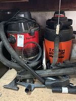 Two shop vacuums side by side on wooden shelf with hoses and multiple attachments in front and around them
