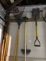 Photo showing two yellow-handled metal-leaf rakes and a white-handled half-moon edger hanging on the wall alongside shovels.