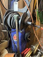Blue Simoniz pressure washer with hose reel and hose coiled, showing the unit in storage with surrounding items.