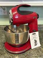 Red Sunbeam MixMaster Series stand mixer with stainless steel bowl on countertop with power outlet in background. User manual placed against bowl.