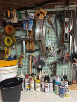 Wide shot showing various hand saws, clamps, paint cans, brushes, and tools hung on pegboard with pail and cans on floor