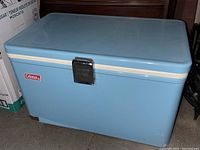 Front view of blue vintage Coleman cooler with white trim and metal latch. Shows peeling finish on bottom edge.