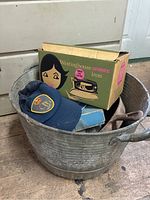 Galvanized metal bin containing a cast iron pan, two hats, a small blue box, and a vintage Westinghouse iron box.