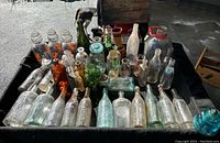 Overview of more than three dozen assorted antique glass bottles in clear, green, and amber shades, some labeled, arranged on a black cloth.