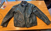 Green leather jacket laid flat on a wooden surface showing front and sleeves in natural light.