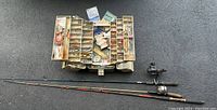 Full view of bifold tacklebox opened showing many compartments filled with assorted fishing lures, sinkers, and accessories, along with three fishing rods laid beneath it.