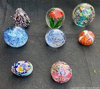 Full view of all seven glass paperweights and one glass egg showing different shapes, colors, and designs.
