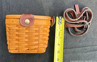 Longaberger wooden basket next to measuring tape showing approximately 5.5 inch height with detached leather strap coiled beside it.