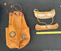 Native American style leather satchel with decorative stitching and brass studs alongside two vintage birchbark canoes and measuring tape to show scale.