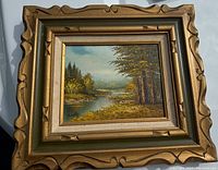 Full view of the framed oil painting showing a river scene with trees and foliage, signed by the artist.
