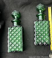 Two green glass decanters, approximately 10 inches tall, square base with diamond texture and round stoppers, shown side by side.