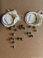 Two silver Pandora bracelets with heart clasps inside white branded pouches, spread on a surface with an assortment of 20 charms made from Murano glass, crystals, and silver accents in various colors and designs.