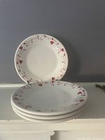 Four white ceramic plates stacked, showing the red heart and vine motif along the rim.
