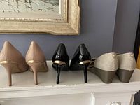 Rear view of three pairs of shoes: nude leather heels, black patent leather heels with gold accents, and beige fabric wedge sandals.