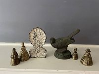 Image showing five brass bells shaped like women in dresses and a metal bird-shaped door stopper on a light-colored surface against a grey wall.