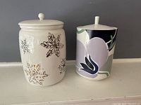 Two ceramic cookie jars side by side against a grey background, showing a white jar with gold snowflakes and a multicolor abstract floral jar.