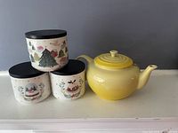 Yellow Le Creuset teapot displayed with three Lapland by Maxwell canisters featuring winter holiday designs.