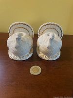 Front view of pair of white ceramic turkey-shaped candle holders with gold accents and a coin for size reference.