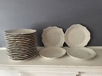 Stack and spread view of Lenox French Perle plates, showing 23 dinner plates stacked and 4 salad/dessert plates in front, all white with scalloped edges and raised beaded snowflake/floral design.
