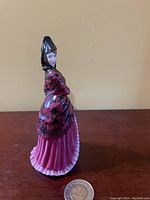 Side view of Royal Doulton Mantilla figurine showing detailed shawl and pink dress with ruffled edges, placed next to a coin for scale.