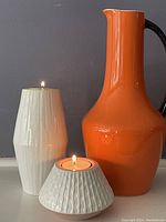 Orange water jug with black handle next to two white ceramic tea light holders, showing relative sizes and shapes